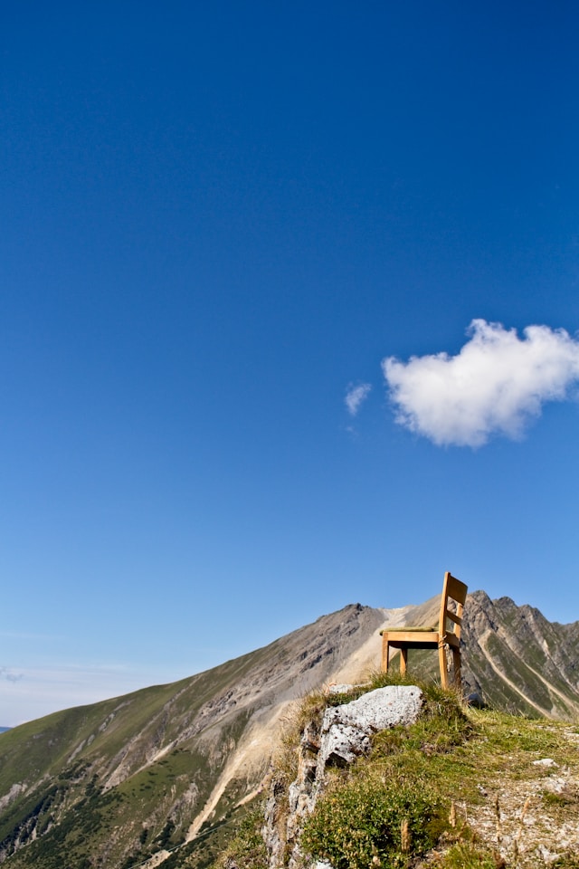 A lone wooden chair at an overlook on a mountain trail under a clear blue sky with a single white cloud.