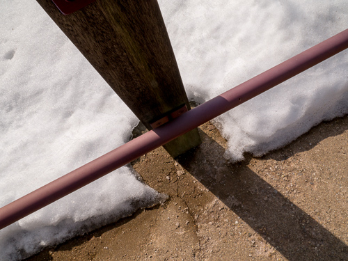 Close-up of a wooden post and a red metal bar in snow with the post casting a shadow on the ground beyond the snow.