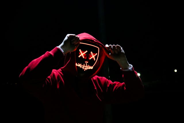 Person in a red hoodie pulling up the hood while wearing an illuminated LED mask featuring X-shaped eyes and a stitched mouth, standing against a dark background.