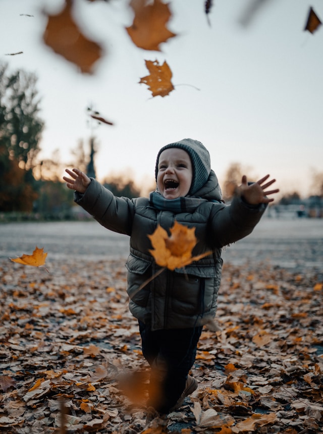 A young child in a coat and hat laughs with arms outstretched as autumn leaves fall and swirl around them in a park.