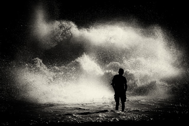 Silhouetted person standing before large breaking ocean waves.