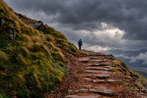 A lone hiker walking up a rocky mountain path through grassy slopes and mist.
