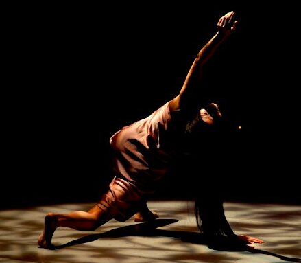A dancer in a low, extended pose under a spotlight against a dark background