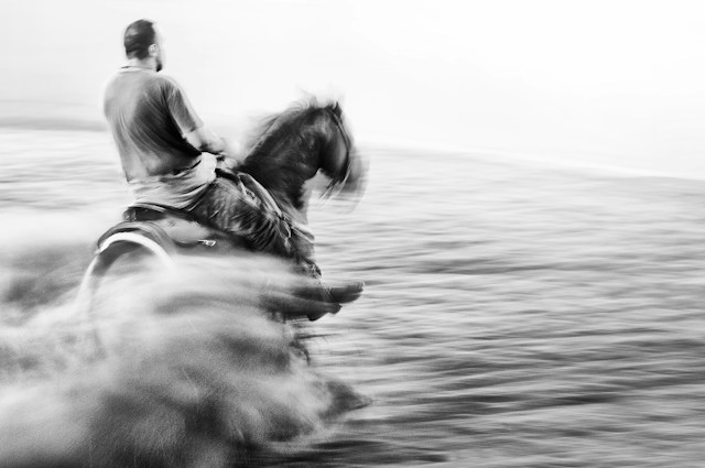 A rider on horseback moving through surf in a blurred black-and-white scene