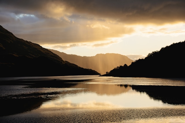 Light breaking through clouds over a mountain lake, reflected in the water between dark slopes.